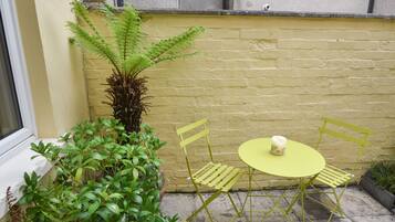 A small outdoor seating area with a table and chairs, surrounded by plants and a yellow wall.