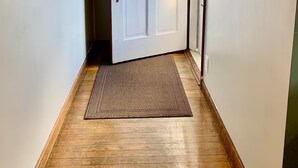 A hallway with a white door, a wooden floor, a doormat, a wall-mounted light, and a framed picture.