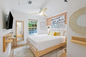 A bedroom with a bed, a desk, a chair, a television, a ceiling fan, a window, and a wall with a framed picture.