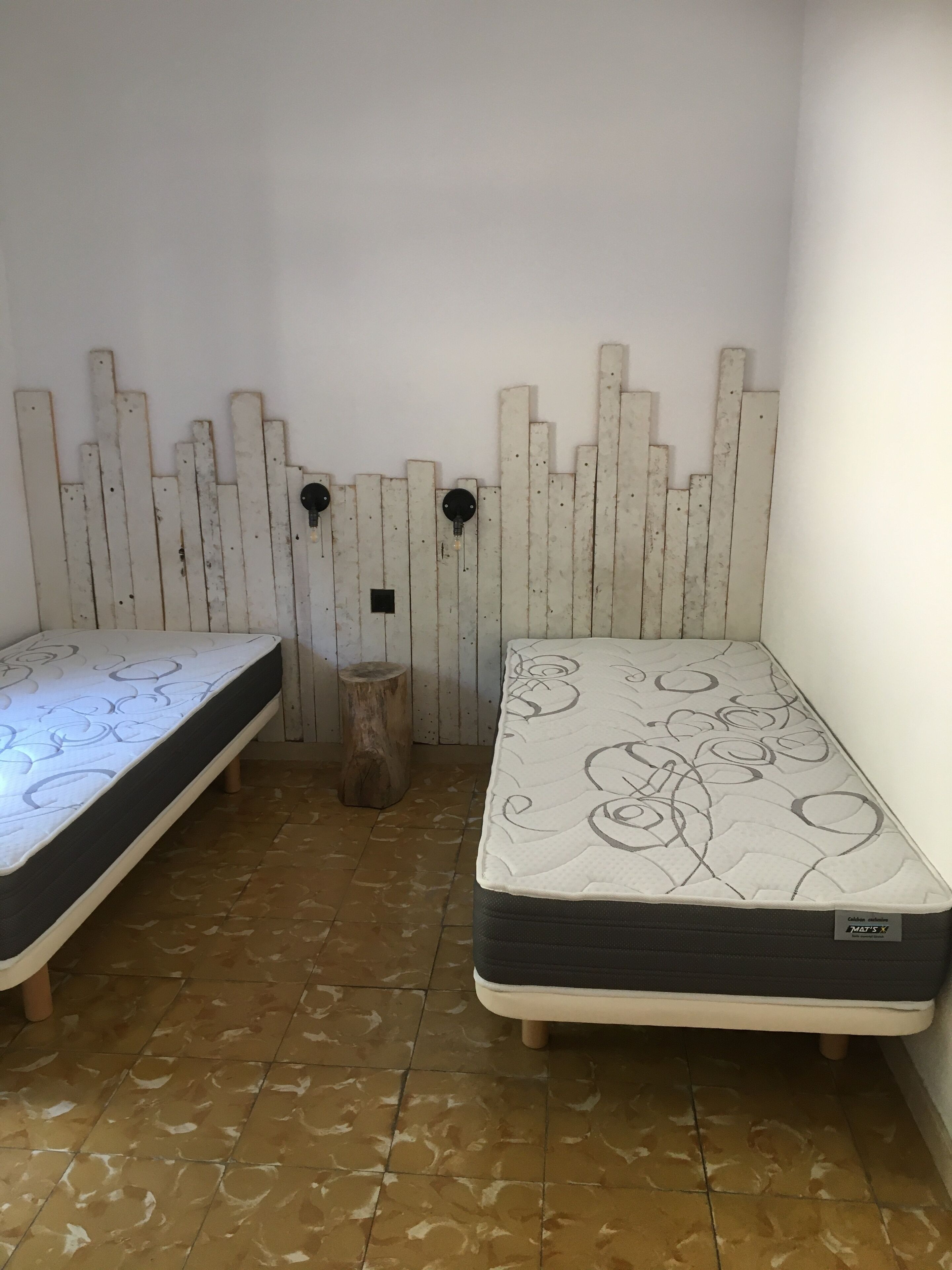 Een kleine kamer met twee enkele bedden, een houten hoofdeinde, een klein krukje en een betegelde vloer.