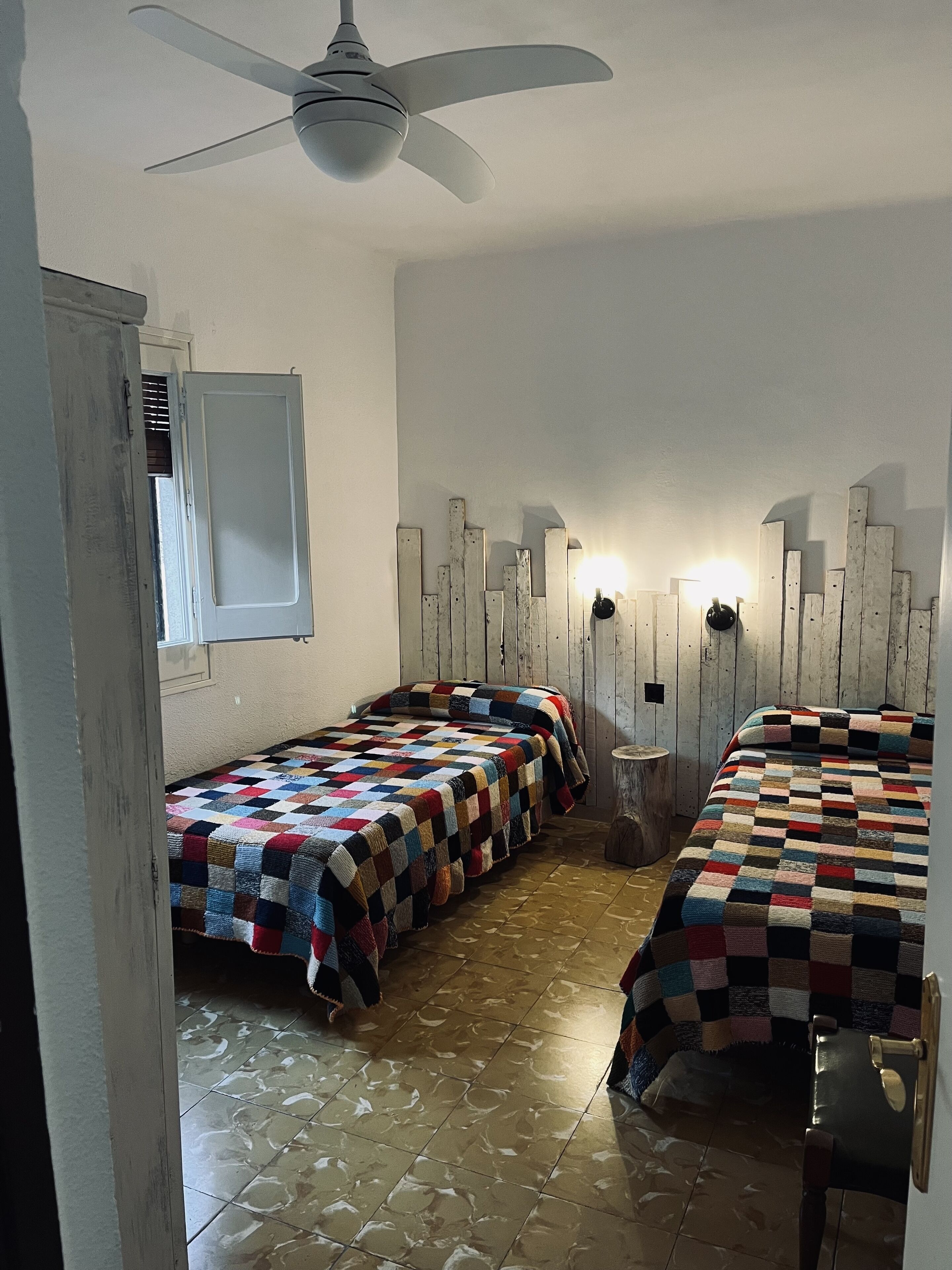 A room with two beds, a ceiling fan, a wall-mounted light, and a window with blinds.