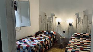 A room with two beds, a ceiling fan, a wall-mounted light, and a window with blinds.