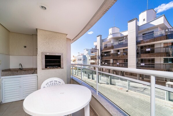 A balcony with a white table and chairs, a small window with a grill, and a view of multi-story buildings.