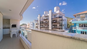 Un balcón con vistas a un edificio de varios pisos, una pequeña mesa con sillas y una ventana con vista al mar.