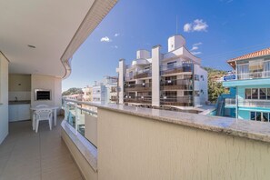 A balcony with a view of a multi-story building, a small table with chairs, and a window with a view of the sea.