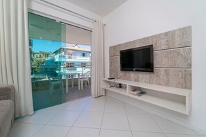A modern living room with a flat-screen TV mounted on a wooden panel wall, a glass door leading to a balcony, and a white tiled floor.