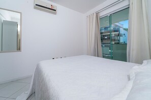 A hotel room with a bed, a window with curtains, a wall-mounted air conditioner, and a view of the hotel exterior.