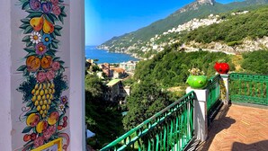 A balcony with a colorful fruit mural, a green railing, and a view of a coastal town with a mountain in the background.