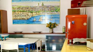 A kitchen with a tiled backsplash, a red refrigerator, a dining table with chairs, and a painting of a coastal town.