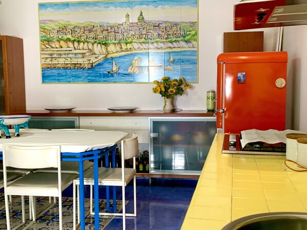 A kitchen with a tiled backsplash, a red refrigerator, a dining table with chairs, and a painting of a coastal town.