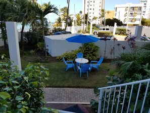 Un patio con sillas azules y una mesa bajo un paraguas azul, rodeado de follaje y una pared blanca.