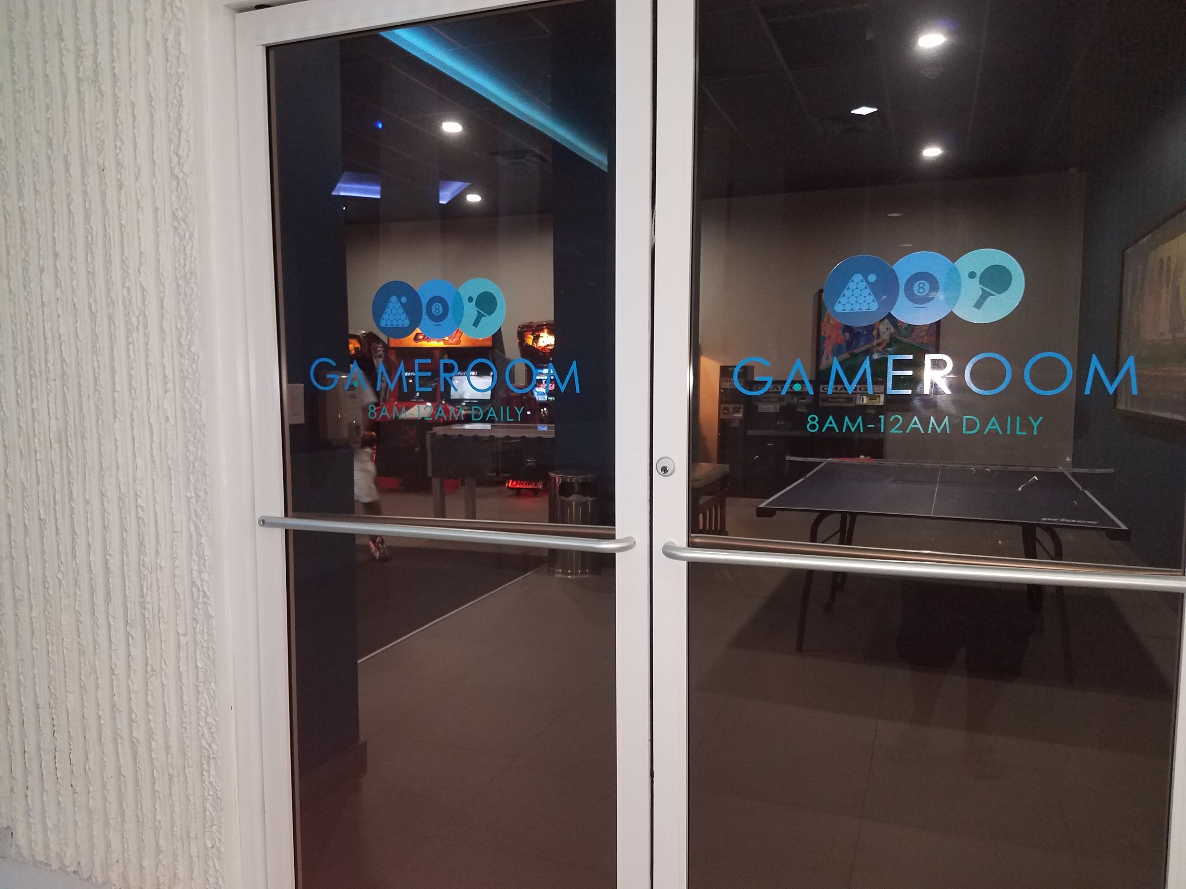 A glass door entrance to a gaming room with a sign indicating it is open daily from 8 AM to 12 AM.