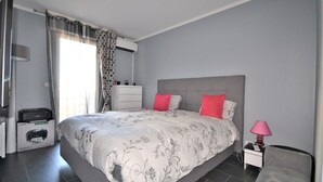 Ein Schlafzimmer mit Bett, Nachttisch, Sessel, Lampe, Schreibtisch mit Drucker und Fenster mit Vorhängen.