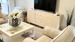 Una sala de estar moderna con un sofá, una mesa de centro con un florero, un televisor sobre un mueble bajo y una ventana con cortinas.