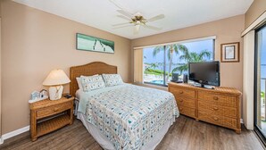 A bedroom with a bed, a television, a nightstand, a lamp, and a window with a view of palm trees and a pool. - Waterfront 2BR/2BA condo with pool, hot tub, balcony, and Wi-Fi. (St. Petersburg)