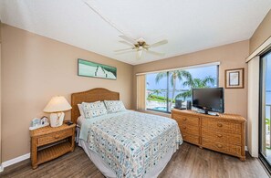 Un dormitorio con cama, televisión, mesita de noche, lámpara y ventana con vistas a palmeras y piscina.