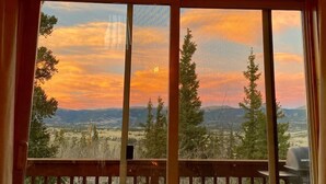 Ein Zimmer mit großen Fenstern und Blick auf einen Sonnenuntergang über einer Berglandschaft.
