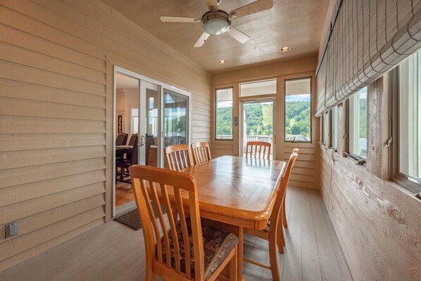 A dining area with a wooden table and chairs, large windows with a view of greenery, and a ceiling fan.