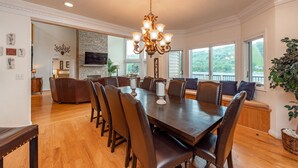 Uma sala de jantar com uma grande mesa de madeira, cadeiras de couro escuro e um candelabro. Há uma lareira de tijolos, uma televisão e vista para o exterior através de amplas janelas.