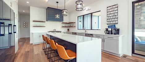A modern kitchen with a large island, bar stools, pendant lighting, and a coffee station.