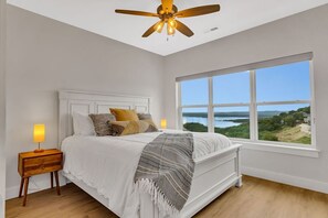 Een slaapkamer met een groot raam, een bed met witte lakens, een houten nachtkastje en een plafondventilator.