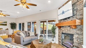 Un salón espacioso con chimenea de piedra, ventilador de techo y un ventanal grande con vistas a un jardín.