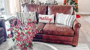 Een woonkamer met een bruine leren bank, een glazen salontafel met een rood bloemarrangement, en een kerstboom op de achtergrond.