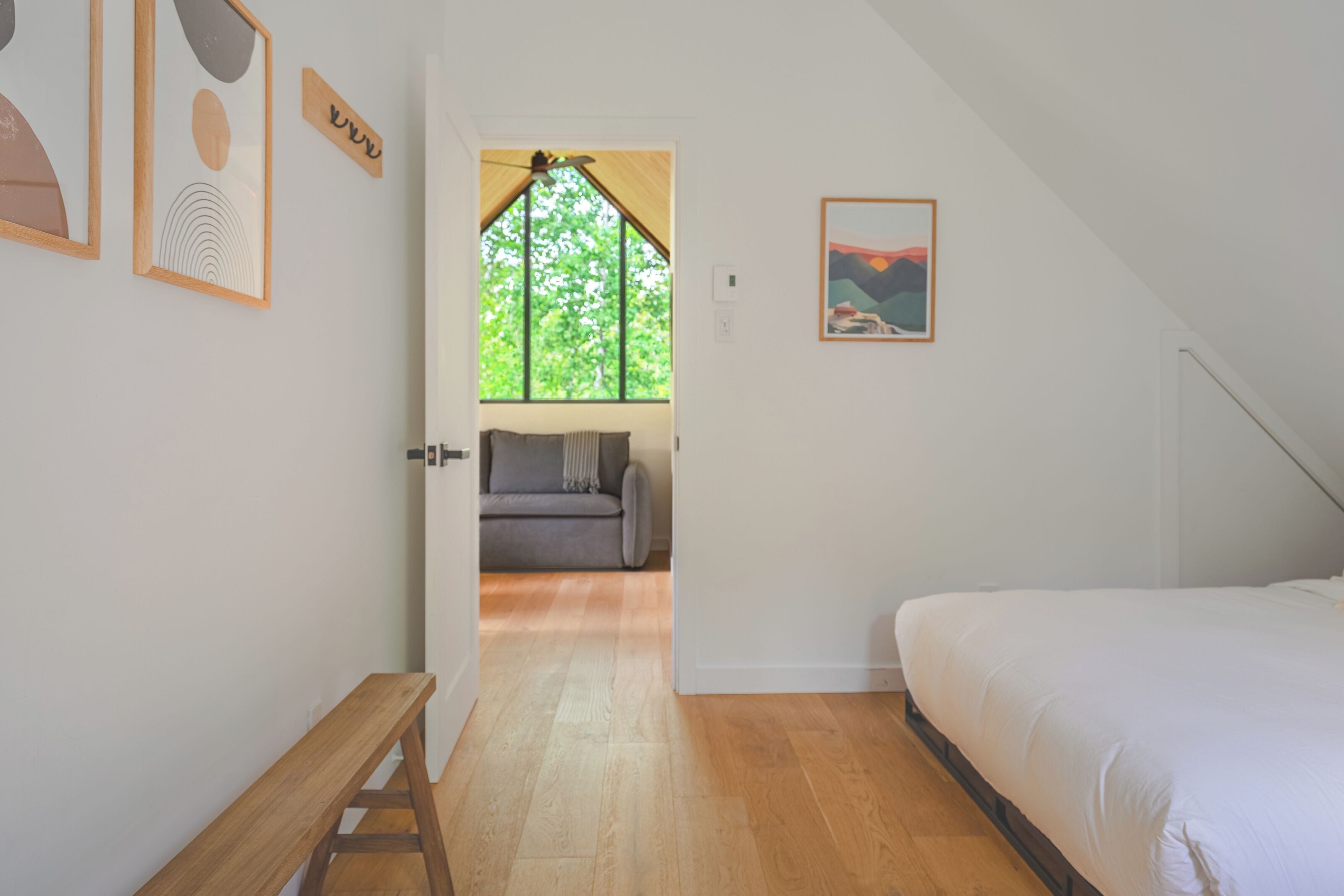 A bedroom with a wooden floor, a bed, a bench, a sofa, and a window with a view of trees.