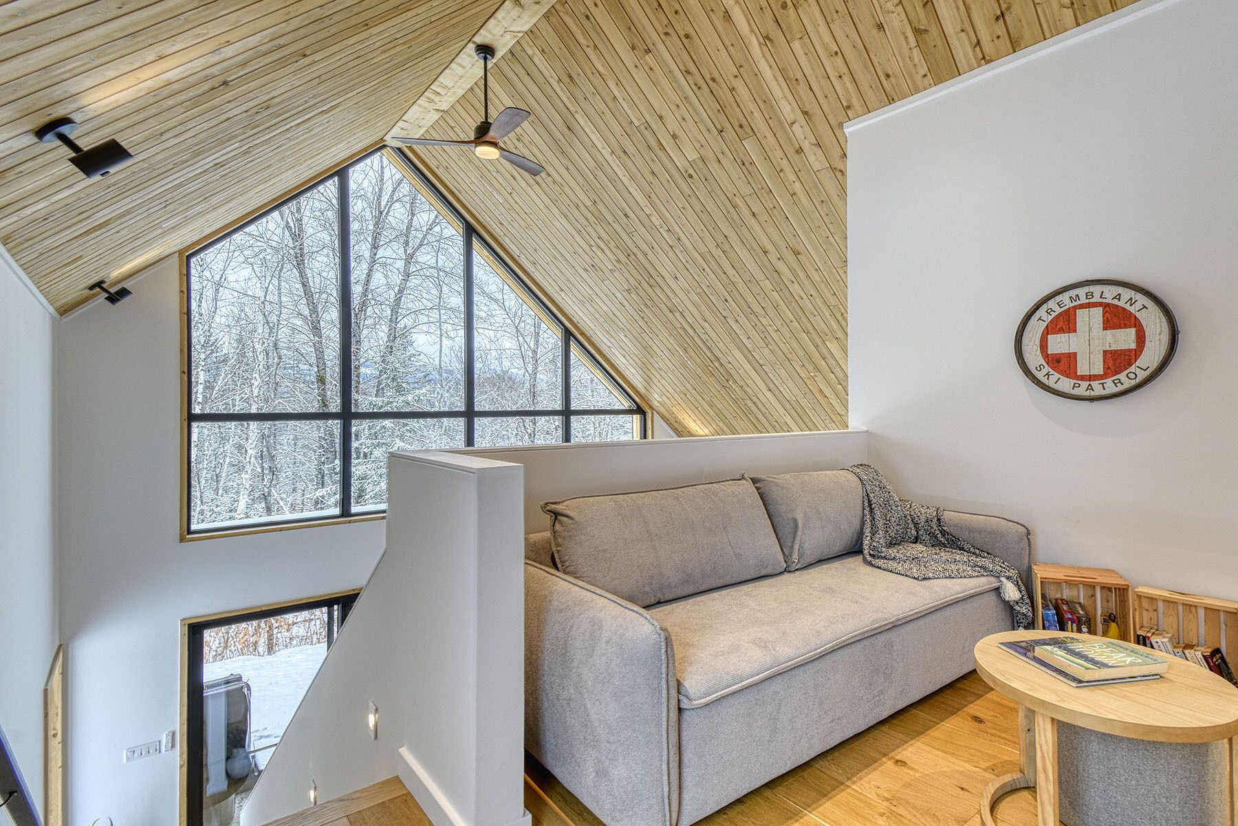 A cozy room with a wooden ceiling, a grey sofa, a small table, and a large window offering a view of snow-covered trees.