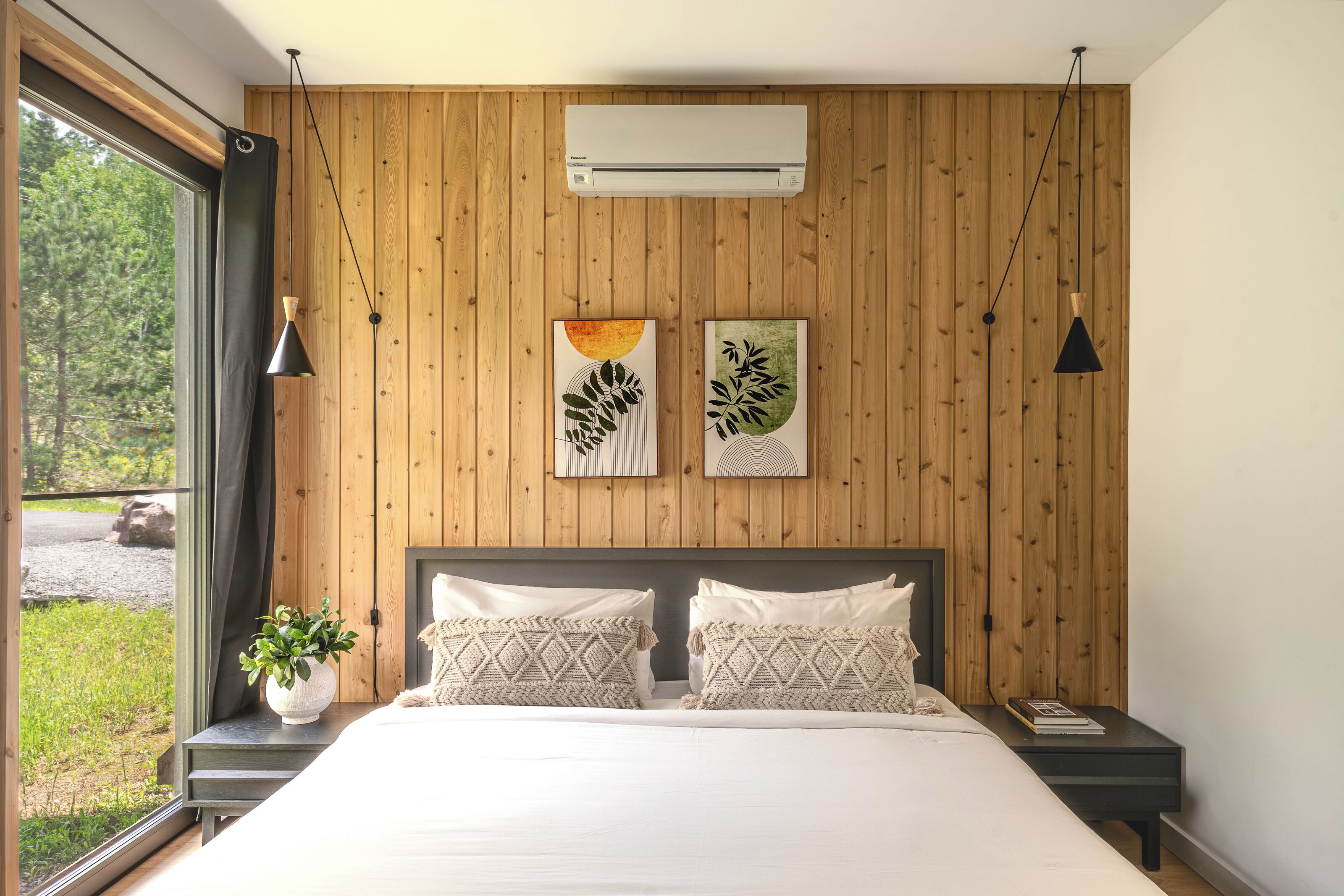 Un dormitorio con pared de paneles de madera, una cama grande con almohadones estampados, una mesita de noche con una planta y obras de arte sobre la cama.