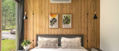 Un dormitorio con pared de paneles de madera, una cama grande con almohadones estampados, una mesita de noche con una planta y obras de arte sobre la cama.