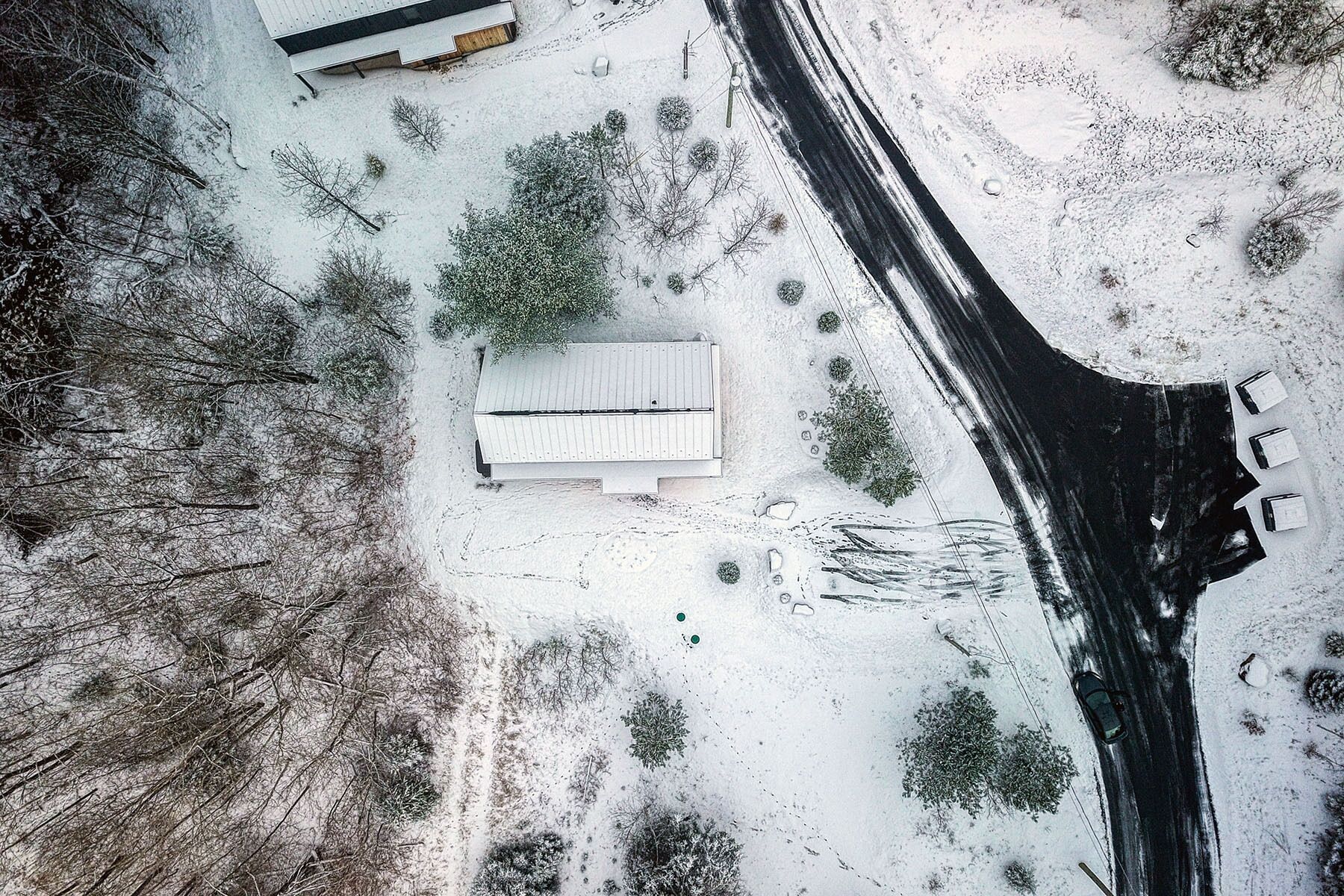 An aerial view of a snow-covered landscape with a road, a building with a sloped roof, and sparse vegetation.