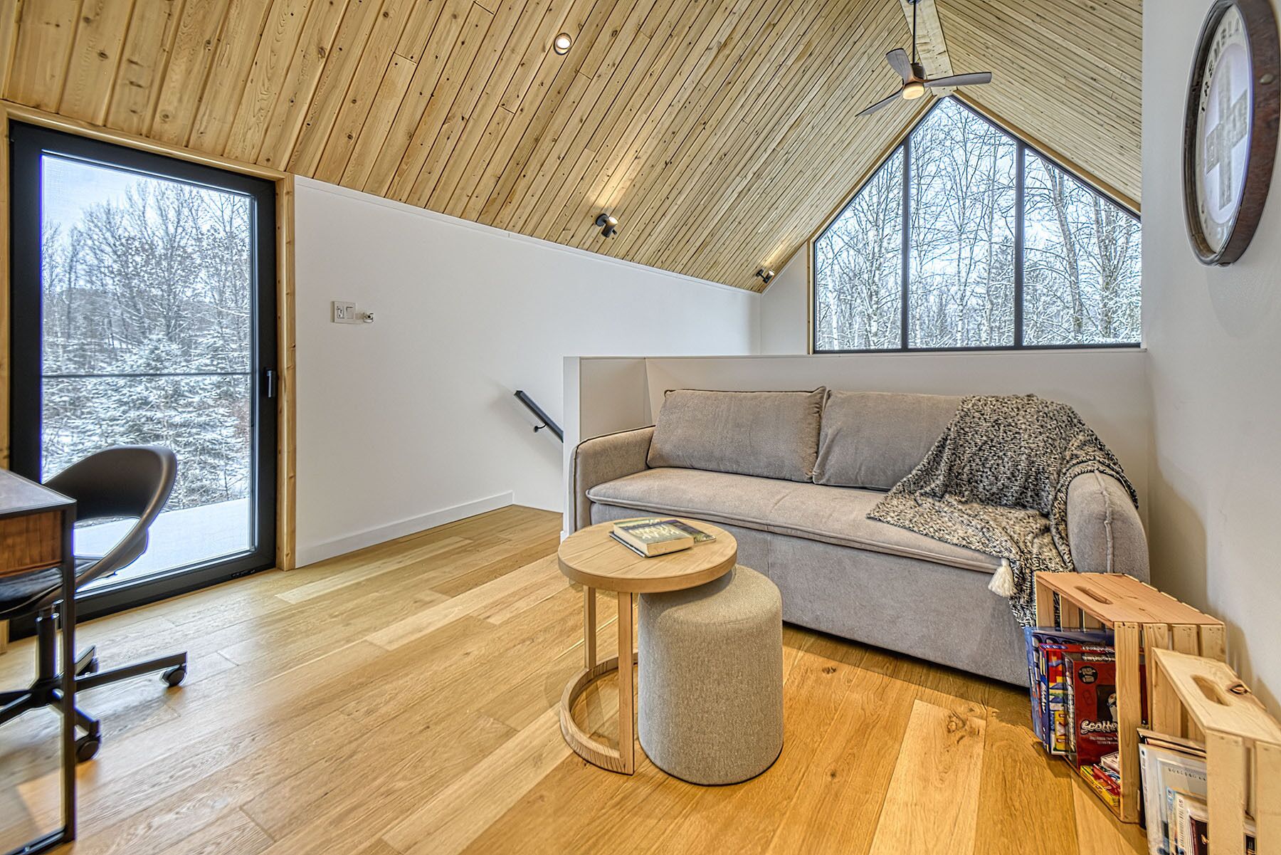 Un salon moderne avec un plafond en bois, un canapé gris, un pouf rond et une vue sur un paysage enneigé par de grandes fenêtres.