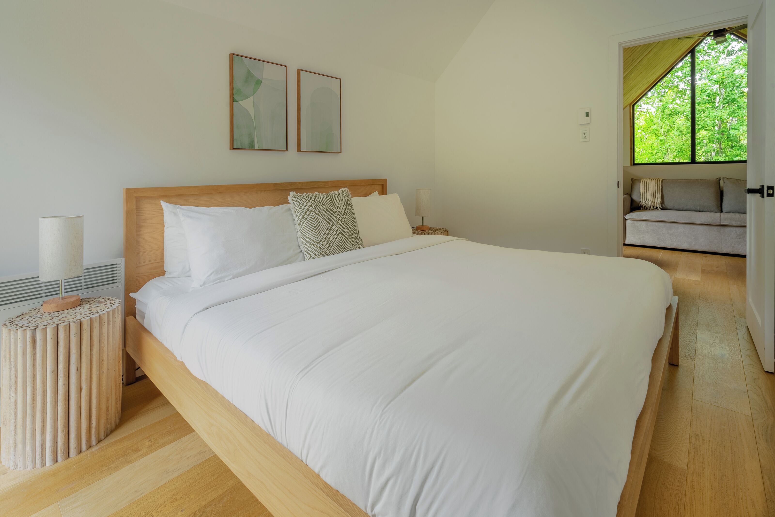 A bedroom with a large bed, a bedside table with a lamp, and a view of a green outdoor area.
