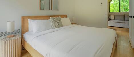 A bedroom with a large bed, a bedside table with a lamp, and a view of a green outdoor area.