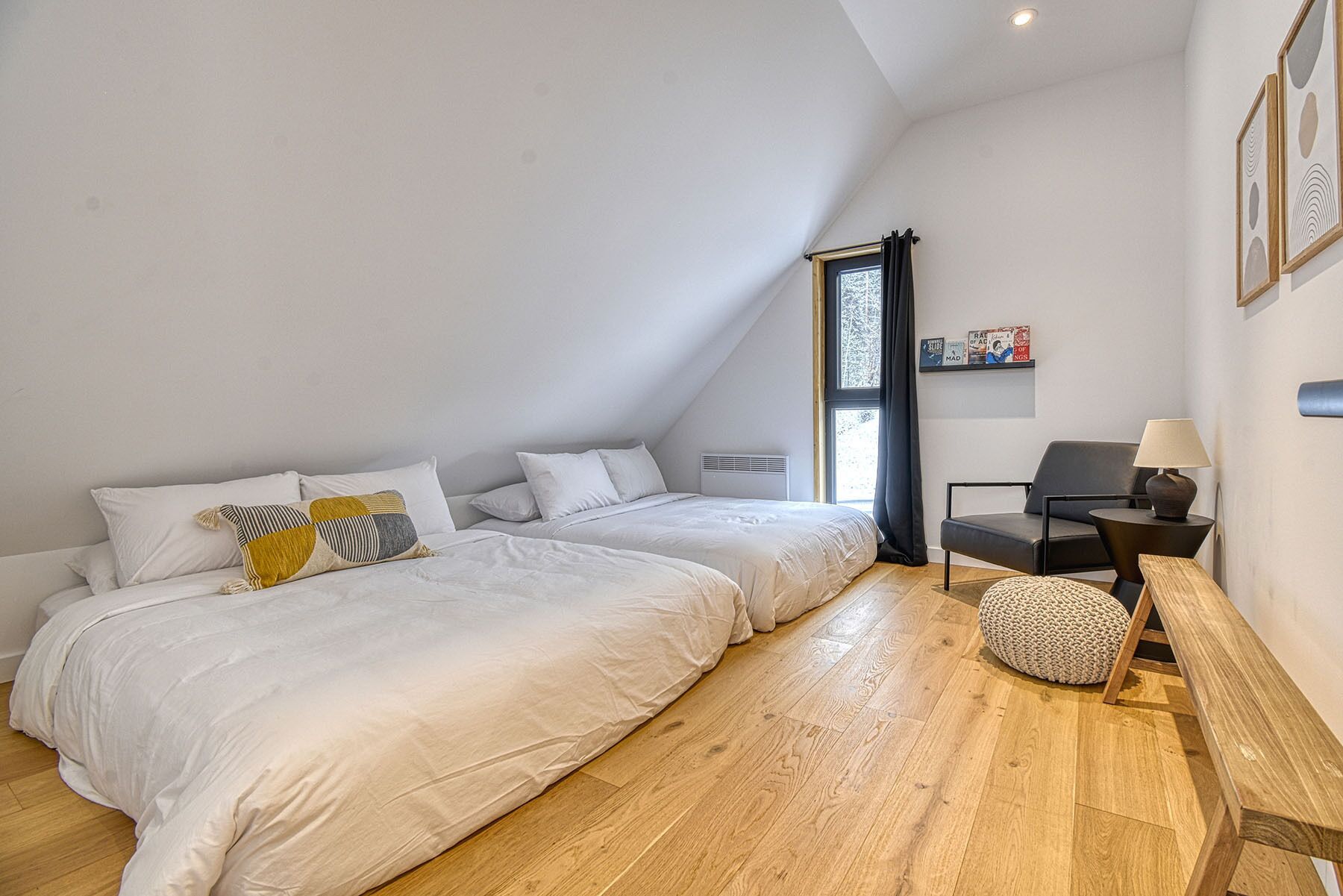 Een slaapkamer met twee bedden, een raam, een stoel, een lamp en een plank met boeken.
