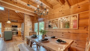 Ein Holzblockhaus-Interieur mit einem Esstisch für vier Personen, einer Küche mit modernen Geräten und einem großen Fenster mit Blick ins Freie.
