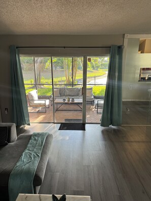 A modern living room with a sofa, a coffee table, and a view through glass doors to an outdoor patio area with a bench and greenery.