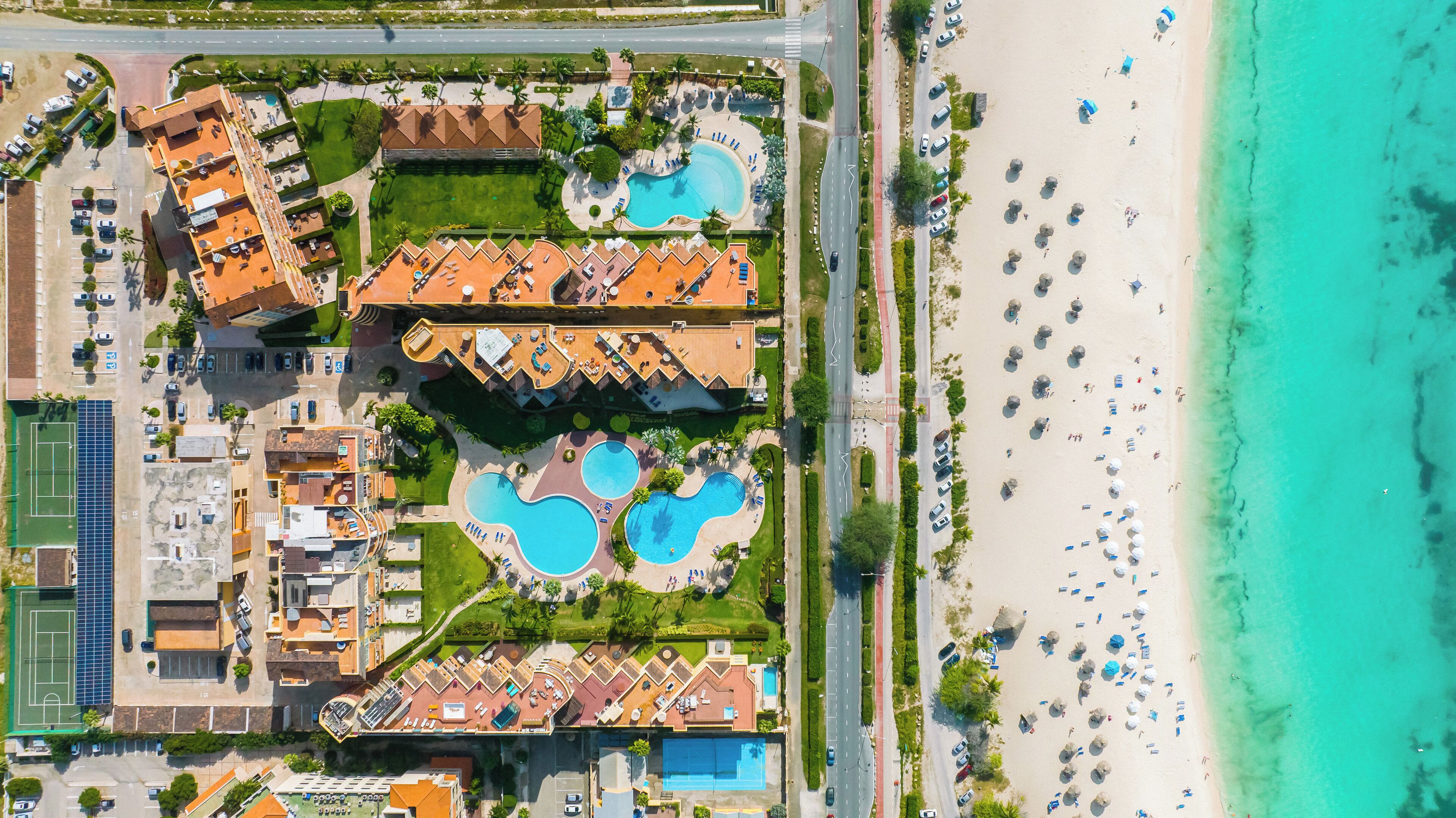 Vista aérea de um resort com várias piscinas, uma praia e uma área de estacionamento.