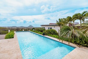 A rectangular swimming pool with clear blue water, surrounded by a tiled deck and lush greenery, including palm trees and various plants.