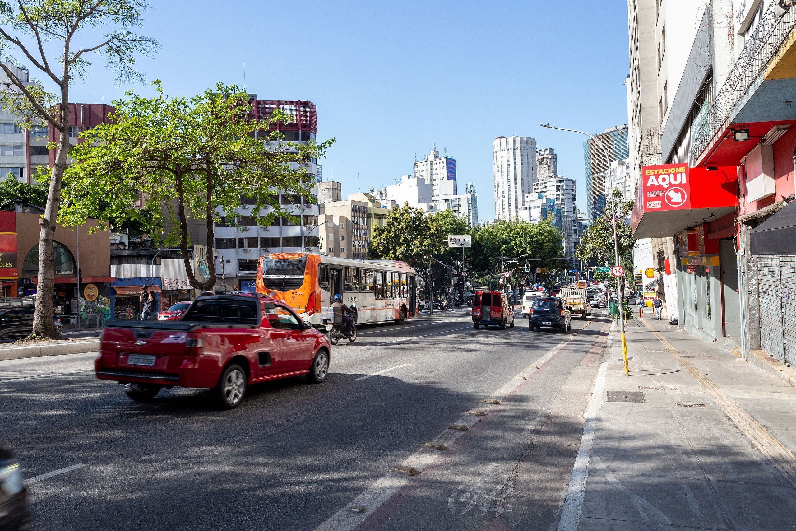 A busy urban street with a red car, a bus, and a van. There are buildings on both sides of the street, including a red building with a sign that says "ESTACIONE AQUI".