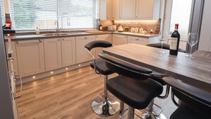 A modern kitchen with wooden flooring, a central island with bar stools, a built-in oven, and a wine bottle on the counter. - Hilltops (Conwy)