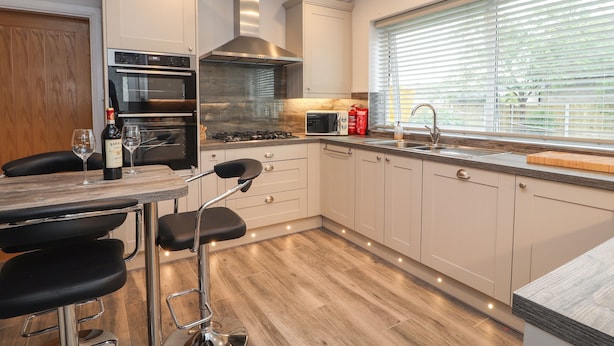 A modern kitchen with a built-in oven, a gas stove, and a stainless steel range hood. There is a bar area with stools and a wine bottle. - Hilltops (Conwy)