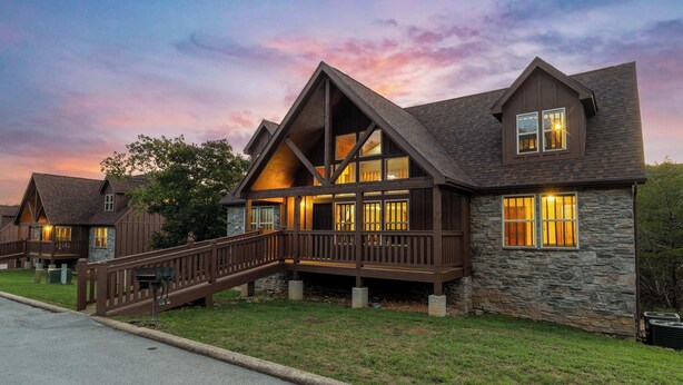 Un grand chalet à deux étages, doté d’une façade en pierre et d’un imposant escalier en bois menant à une terrasse.