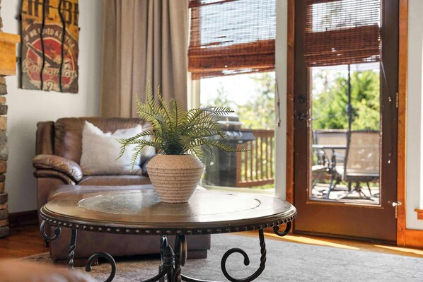 Una sala de estar con un sofá de cuero marrón, una mesa redonda con una planta y una puerta que da a un patio exterior.