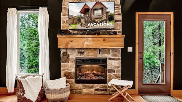 Una chimenea de piedra con un televisor que muestra 'NIGHTLY VACATIONS', una cesta de mimbre con una toalla blanca y una puerta de madera que da a una zona boscosa.