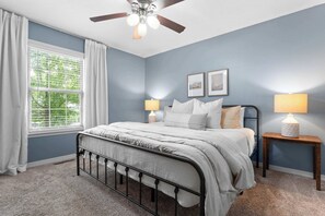 A bedroom with a bed, a ceiling fan, two bedside lamps, a window with curtains, and a wall-mounted picture.