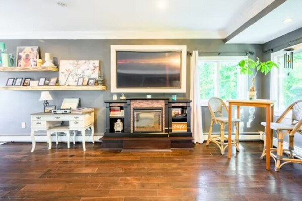 A living room with a fireplace, wooden flooring, a desk, wicker chairs, and a large window with a view of greenery.
