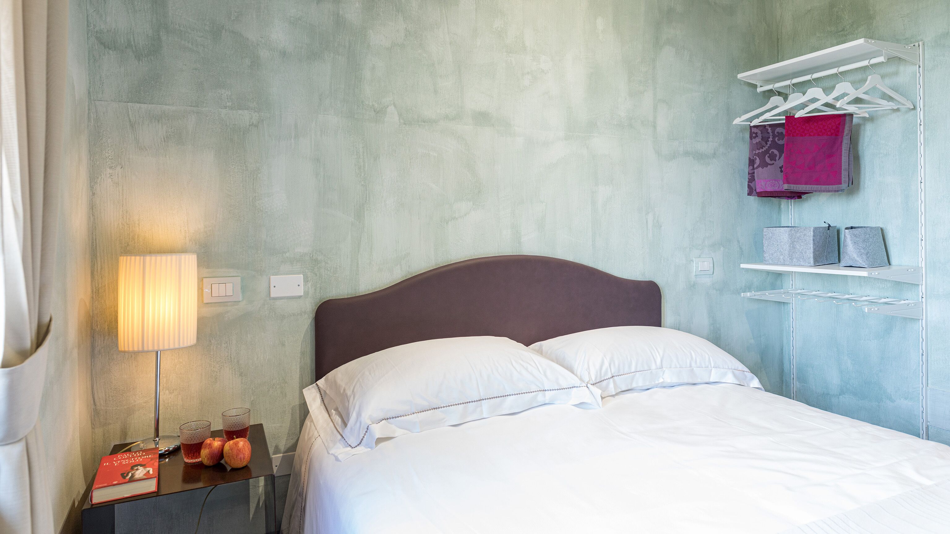 Un lit bien fait dans une pièce dont le mur est texturé, une table de chevet avec une lampe, un livre et deux pommes, ainsi qu’un portant à vêtements avec des cintres et des sacs.
