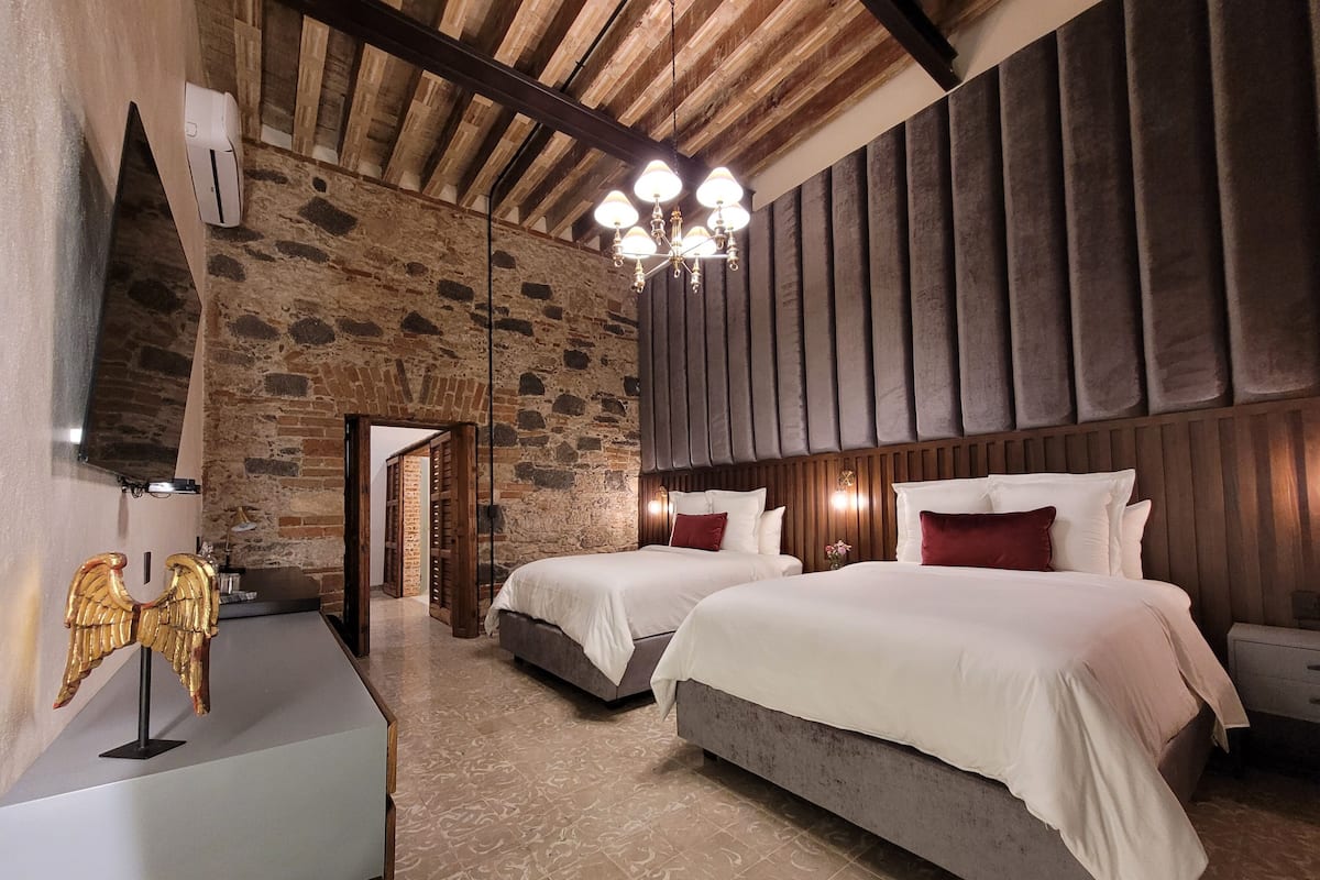 A hotel room with two beds, a stone wall, a wooden ceiling, and a chandelier.